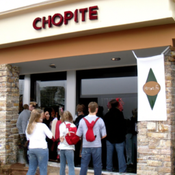 description: A photo of a Chipotle Mexican Grill restaurant, with customers standing in line to order food. The restaurant's logo is visible on the storefront, and a sign advertises a new menu item. The image is taken from an anonymous source and does not contain any identifiable information.